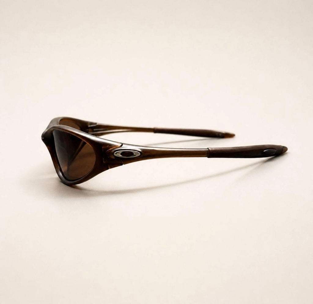 Side view of brown Oakley Minute sunglasses, showing the brown frame, lens and silver oval temple detail.